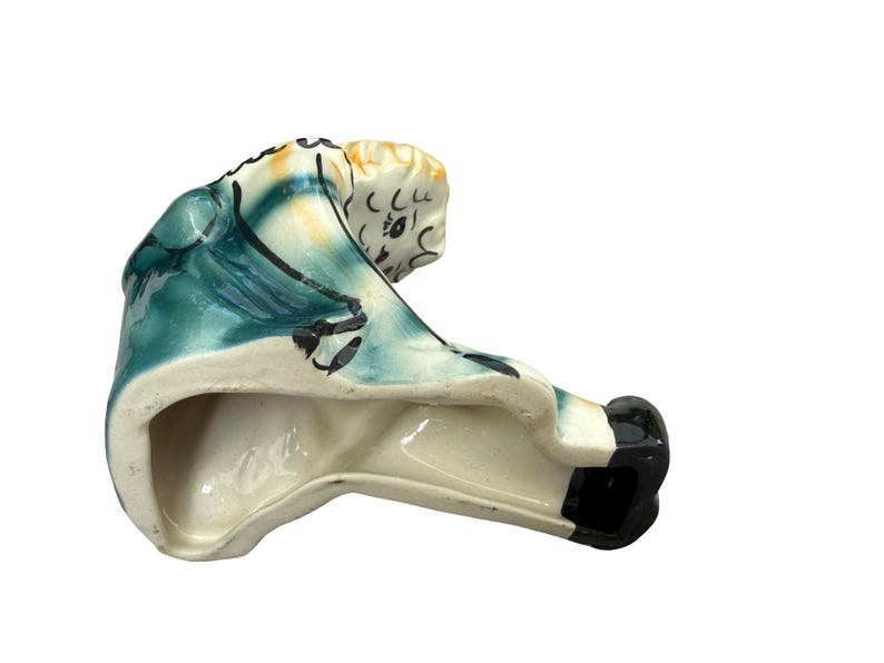May include: A ceramic figurine of a seated figure, painted with teal, cream, and black accents. The figure has a detailed face and is wearing a teal garment. The base is unglazed, revealing the ceramic material.