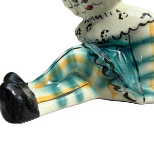 May include: A ceramic figurine of a seated clown with a white head and a ruffled collar. The clown's outfit features a teal and yellow plaid pattern. The clown's shoes are black.