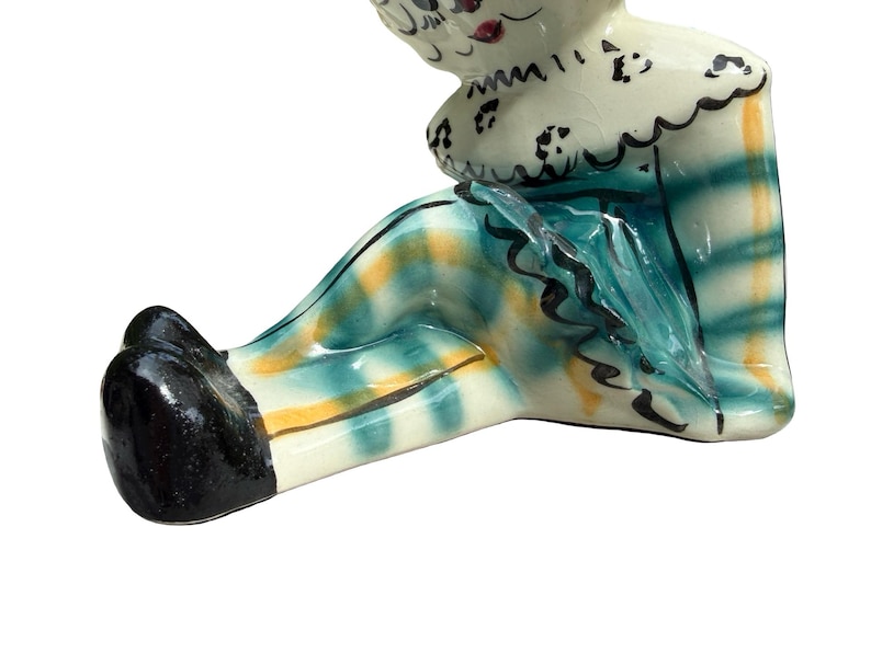May include: A ceramic figurine of a seated clown with a white head and a ruffled collar. The clown's outfit features a teal and yellow plaid pattern. The clown's shoes are black.