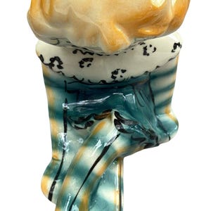 May include: A ceramic figurine of a chef, featuring a yellow hat and a blue and yellow striped uniform. The figurine has a black shoe and a white collar with black detailing. The chef's hat is a light yellow color.