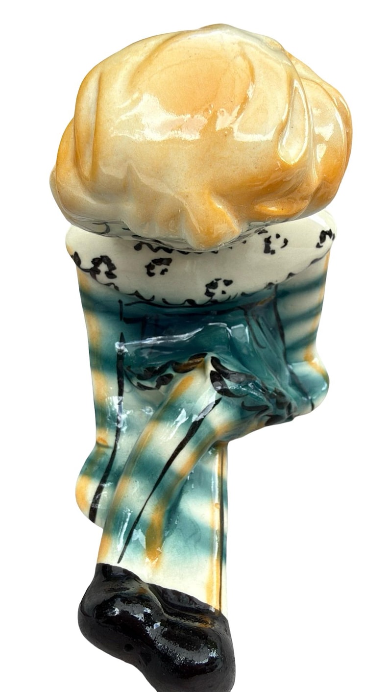 May include: A ceramic figurine of a chef, featuring a yellow hat and a blue and yellow striped uniform. The figurine has a black shoe and a white collar with black detailing. The chef's hat is a light yellow color.