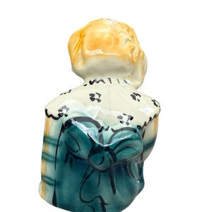 May include: A ceramic figurine of a person, viewed from the back. The figure has blonde hair and is wearing a teal dress with a large bow. The dress has a plaid pattern on the sides. The collar is white with black floral accents.