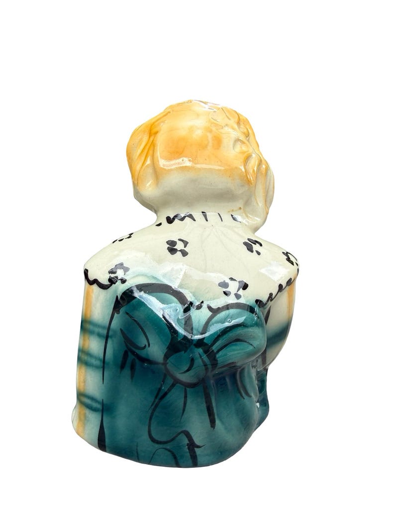 May include: A ceramic figurine of a person, viewed from the back. The figure has blonde hair and is wearing a teal dress with a large bow. The dress has a plaid pattern on the sides. The collar is white with black floral accents.