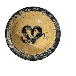 May include: A round, ceramic bowl with a speckled tan interior and a dark blue rim. The bowl features a hand-painted heart design in dark blue in the center. The rim has a textured, speckled appearance.