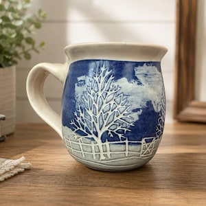 May include: A ceramic mug with a blue and white winter scene design. The mug features a white tree and fence design against a blue background. The handle and rim are a cream color. The mug is sitting on a wooden surface.