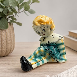 May include: A vintage ceramic figurine of a seated doll with a yellow and orange head, and a plaid green, yellow, and blue outfit. The doll has black shoes and a white collar with black detailing. The figurine is on a wooden surface.