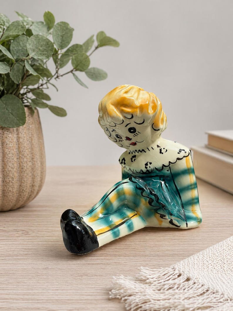May include: A vintage ceramic figurine of a seated doll with a yellow and orange head, and a plaid green, yellow, and blue outfit. The doll has black shoes and a white collar with black detailing. The figurine is on a wooden surface.