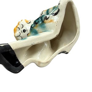 May include: A vintage ceramic planter in the shape of a woman reclining. The planter is painted with a cream base, black accents, and colorful details on the figure's head and clothing. The interior is unglazed.