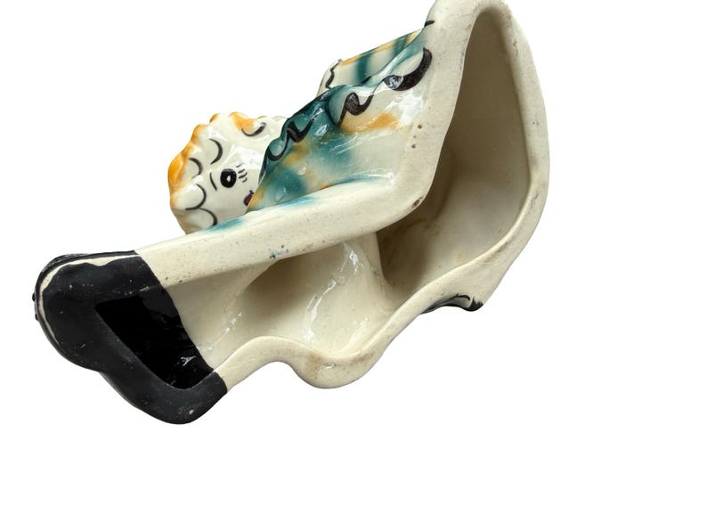 May include: A vintage ceramic planter in the shape of a woman reclining. The planter is painted with a cream base, black accents, and colorful details on the figure's head and clothing. The interior is unglazed.