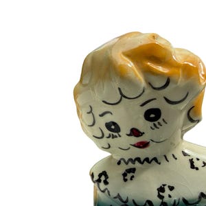 May include: A vintage ceramic figurine of a young girl with blonde hair and a blue dress. The figurine has black painted eyes, eyebrows, and a red mouth. The girl's dress has a scalloped collar and black floral accents.