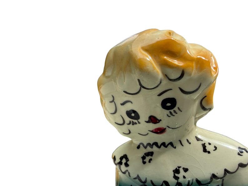 May include: A vintage ceramic figurine of a young girl with blonde hair and a blue dress. The figurine has black painted eyes, eyebrows, and a red mouth. The girl's dress has a scalloped collar and black floral accents.