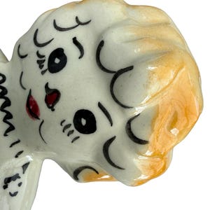 May include: A ceramic figurine head with a white base and orange-yellow hair. The face has black eyes, eyelashes, and eyebrows, with a red mouth. The figurine has a glossy finish and is likely a decorative item.