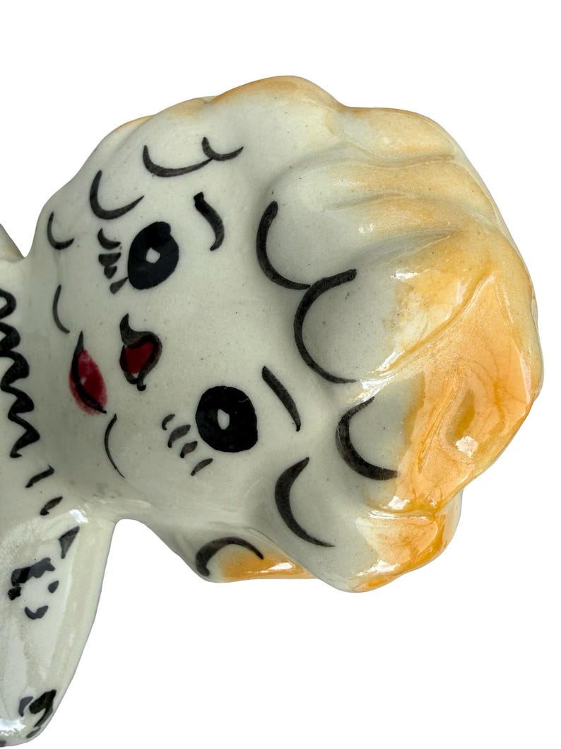 May include: A ceramic figurine head with a white base and orange-yellow hair. The face has black eyes, eyelashes, and eyebrows, with a red mouth. The figurine has a glossy finish and is likely a decorative item.