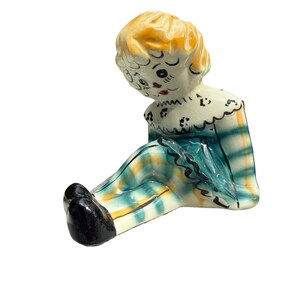 May include: A vintage ceramic figurine of a seated doll with a yellow head and black shoes. The doll wears a white ruffled collar and a plaid outfit in shades of teal, yellow, and white. The doll's face has painted features.