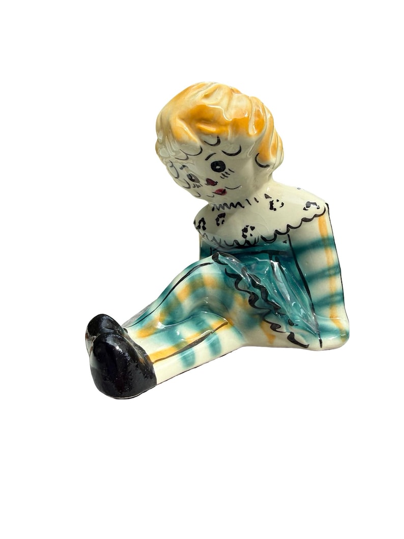 May include: A vintage ceramic figurine of a seated doll with a yellow head and black shoes. The doll wears a white ruffled collar and a plaid outfit in shades of teal, yellow, and white. The doll's face has painted features.