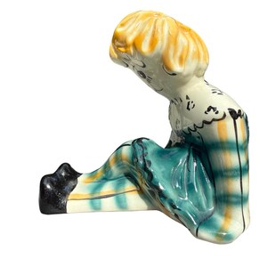 May include: A ceramic figurine of a seated figure with yellow hair and a teal and yellow plaid outfit. The figurine has black shoes and a white base. The figure is looking down.
