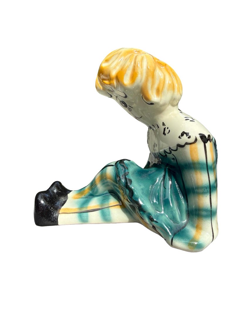 May include: A ceramic figurine of a seated figure with yellow hair and a teal and yellow plaid outfit. The figurine has black shoes and a white base. The figure is looking down.