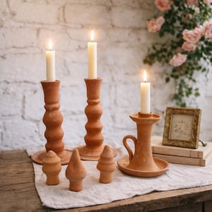 May include: A collection of terracotta candle holders and decorative pieces. The candle holders vary in size and design, with lit white candles. The pieces are arranged on a wooden table with a neutral linen cloth, against a white brick wall.