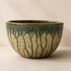 May include: A round ceramic bowl featuring a textured, speckled cream-colored base with a reactive glaze effect. The upper portion of the bowl displays a deep teal blue drip pattern that cascades down the sides, ending in a dark, earthy rim.