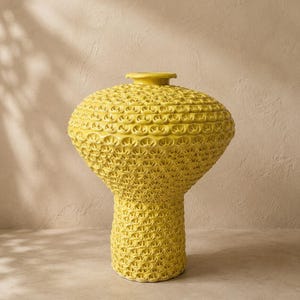 May include: A bright yellow ceramic vase with a wide, rounded upper body and a narrower cylindrical base. The entire surface features a repeating, textured, honeycomb-like pattern of small, raised, semi-circular arches across the exterior.