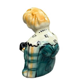 May include: A ceramic figurine of a person with blonde hair, wearing a teal dress with black accents and a white collar with black floral details. The figurine is viewed from the back and slightly to the side.