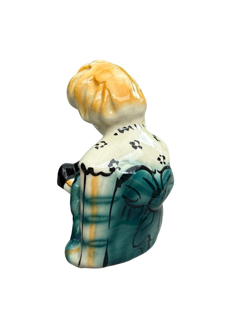 May include: A ceramic figurine of a person with blonde hair, wearing a teal dress with black accents and a white collar with black floral details. The figurine is viewed from the back and slightly to the side.