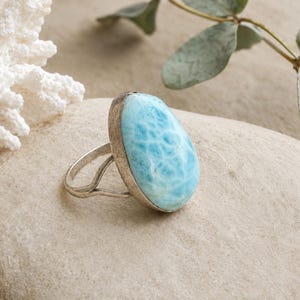 May include: A silver ring featuring a large, oval-shaped, light blue Larimar gemstone with white marbling patterns. The stone is set in a smooth metal bezel, and the band consists of a split-shank design that tapers toward the back of the ring.