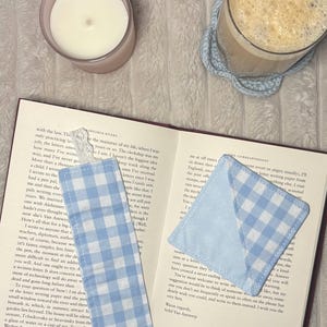 May include: A rectangular fabric bookmark with a light blue and white gingham pattern and a white lace trim at the top, alongside a square corner bookmark featuring the same light blue and white gingham fabric, both resting on an open book.