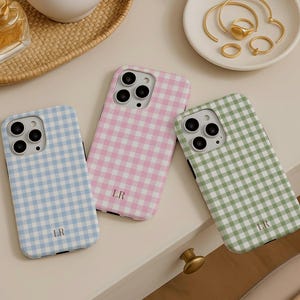 Puede incluir: Tres fundas para smartphone con un patrón de cuadros vichy en azul claro, rosa y verde salvia. Cada funda tiene un recorte para la cámara y las iniciales LR impresas en una fuente pequeña y oscura en la parte inferior central.