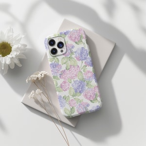 May include: A white phone case featuring a watercolor floral design of pink and purple hydrangeas with green leaves. The case is resting on a white surface with a daisy and dried flowers nearby.