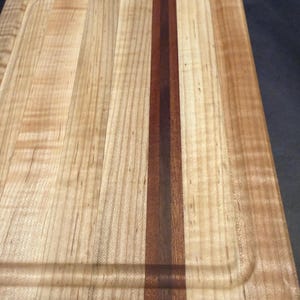 May include: A rectangular wooden cutting board featuring a light-colored wood grain with a prominent, dark brown vertical stripe running through the center. The board includes a recessed juice groove carved along the perimeter of the surface.