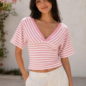 May include: A short-sleeved top featuring a pink and white horizontal striped pattern with a deep V-neckline and a solid pink trim. The garment is cropped at the waist and includes the text suki SEW printed in the upper right corner.