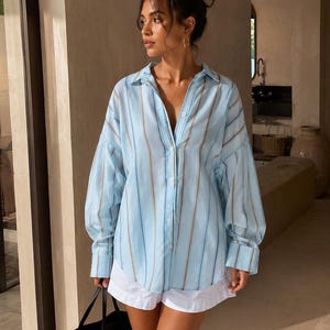 May include: A light blue button-down shirt featuring vertical stripes in shades of white, tan, and darker blue, paired with white shorts and a black and tan woven tote bag. The shirt has a collar, long sleeves with cuffs, and a loose fit.