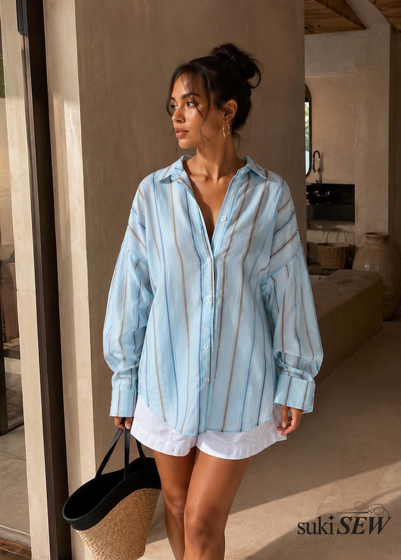 May include: A light blue button-down shirt featuring vertical stripes in shades of white, tan, and darker blue, paired with white shorts and a black and tan woven tote bag. The shirt has a collar, long sleeves with cuffs, and a loose fit.