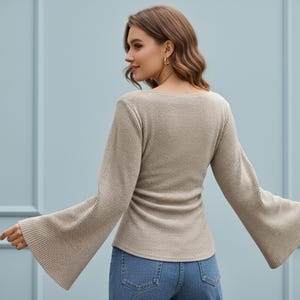 May include: A beige long-sleeved top featuring a ribbed knit texture and wide, flared bell sleeves. The garment has a square neckline and a fitted bodice that extends to the hips, paired with blue denim jeans.