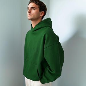 Puede incluir: Una sudadera con capucha de color verde bosque s&oacute;lido que presenta un espacioso bolsillo tipo canguro frontal, mangas largas con pu&ntilde;os acanalados y una capucha estructurada. La prenda est&aacute; confeccionada en tela gruesa y lisa, con pantalones blancos.