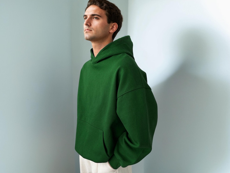 Puede incluir: Una sudadera con capucha de color verde bosque s&oacute;lido que presenta un espacioso bolsillo tipo canguro frontal, mangas largas con pu&ntilde;os acanalados y una capucha estructurada. La prenda est&aacute; confeccionada en tela gruesa y lisa, con pantalones blancos.