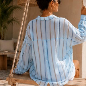 May include: A light blue button-down shirt featuring vertical stripes in shades of blue and tan. The shirt has long sleeves with cuffs, a classic collar, and a vertical row of buttons running down the center of the back. The fabric appears lightweight.