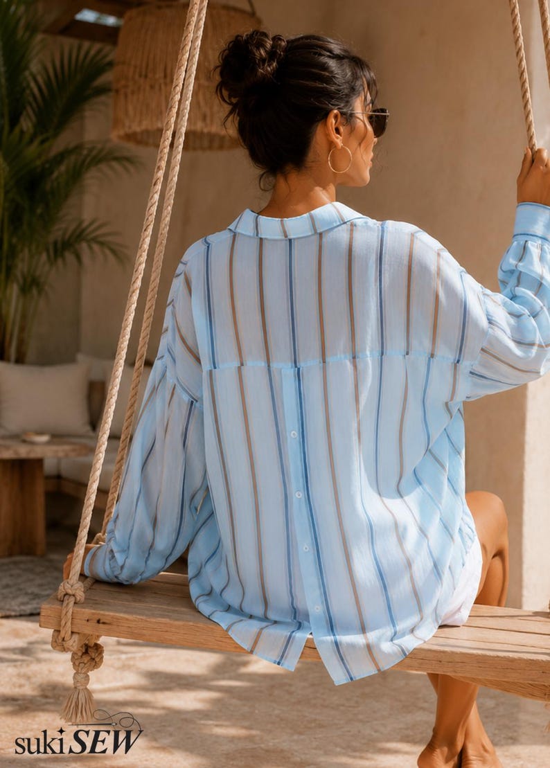 May include: A light blue button-down shirt featuring vertical stripes in shades of blue and tan. The shirt has long sleeves with cuffs, a classic collar, and a vertical row of buttons running down the center of the back. The fabric appears lightweight.