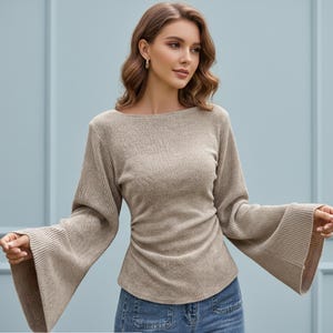 May include: A beige long-sleeved top featuring a ribbed knit texture, a wide boat neckline, and dramatic flared bell sleeves. The garment has a fitted bodice with subtle side ruching at the waist and is paired with blue denim jeans.