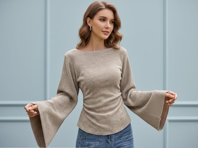May include: A beige long-sleeved top featuring a ribbed knit texture, a wide boat neckline, and dramatic flared bell sleeves. The garment has a fitted bodice with subtle side ruching at the waist and is paired with blue denim jeans.