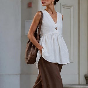 May include: A sleeveless white peplum top with a V-neckline and a single dark brown button closure, paired with loose-fitting dark brown trousers and a textured brown woven shoulder bag. The image includes the text suki SEW in the top left corner.