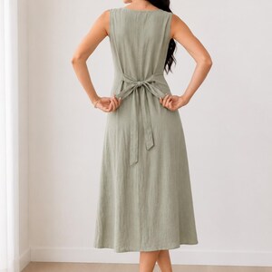Peut inclure: Une robe midi sans manches vert sauge avec une encolure ronde et un lien &agrave; nouer &agrave; la taille dans le dos. La robe pr&eacute;sente un design simple et uni avec une jupe &eacute;vas&eacute;e qui tombe sous les genoux. Le texte suki SEW est visible en haut &agrave; droite.
