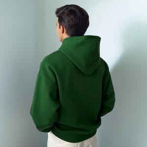 Puede incluir: Una sudadera con capucha de color verde bosque s&oacute;lido con mangas largas, capucha sin cordones y dobladillo acanalado. La prenda se muestra desde la parte posterior, exhibiendo un dise&ntilde;o liso sin patrones con un ajuste relajado y textura mate.