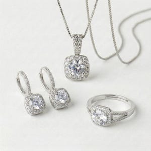 May include: A jewelry set featuring a silver necklace with a square pendant, matching earrings, and a ring. Each piece is adorned with a large, clear, round-cut stone surrounded by smaller stones. The set is displayed on a white surface.