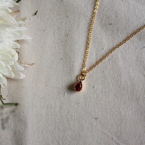 May include: A delicate gold chain necklace featuring a small, teardrop-shaped pendant with a deep red gemstone. The pendant is framed by a thin, textured gold border, and the chain consists of small, uniform oval links that catch the light.