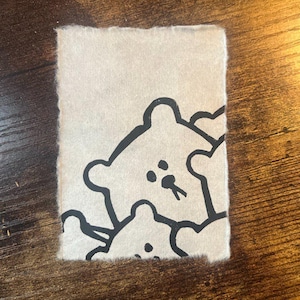 May include: A rectangular piece of off-white paper with a black line drawing of three bear-like figures. The bears have simple features, including small dots for eyes and a small line for a mouth. The paper has a rough, textured edge.