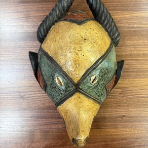 Puede incluir: Una máscara de madera hecha a mano con una cara amarilla, detalles verdes y rojos y cuernos negros. La máscara presenta dos pequeños detalles blancos en forma de concha y probablemente se utiliza con fines decorativos o ceremoniales.