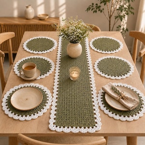 May include: A set of olive green crochet placemats and a matching table runner, each featuring a decorative white scalloped border. The set is arranged on a wooden table with ceramic plates, a tea cup, a vase of flowers, and a small candle.