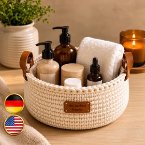 May include: A round, cream-colored crocheted basket with two tan leather handles and a brown leather label reading the perfect looper. The basket contains two pump bottles, two small jars, a dropper bottle, and a folded white towel.